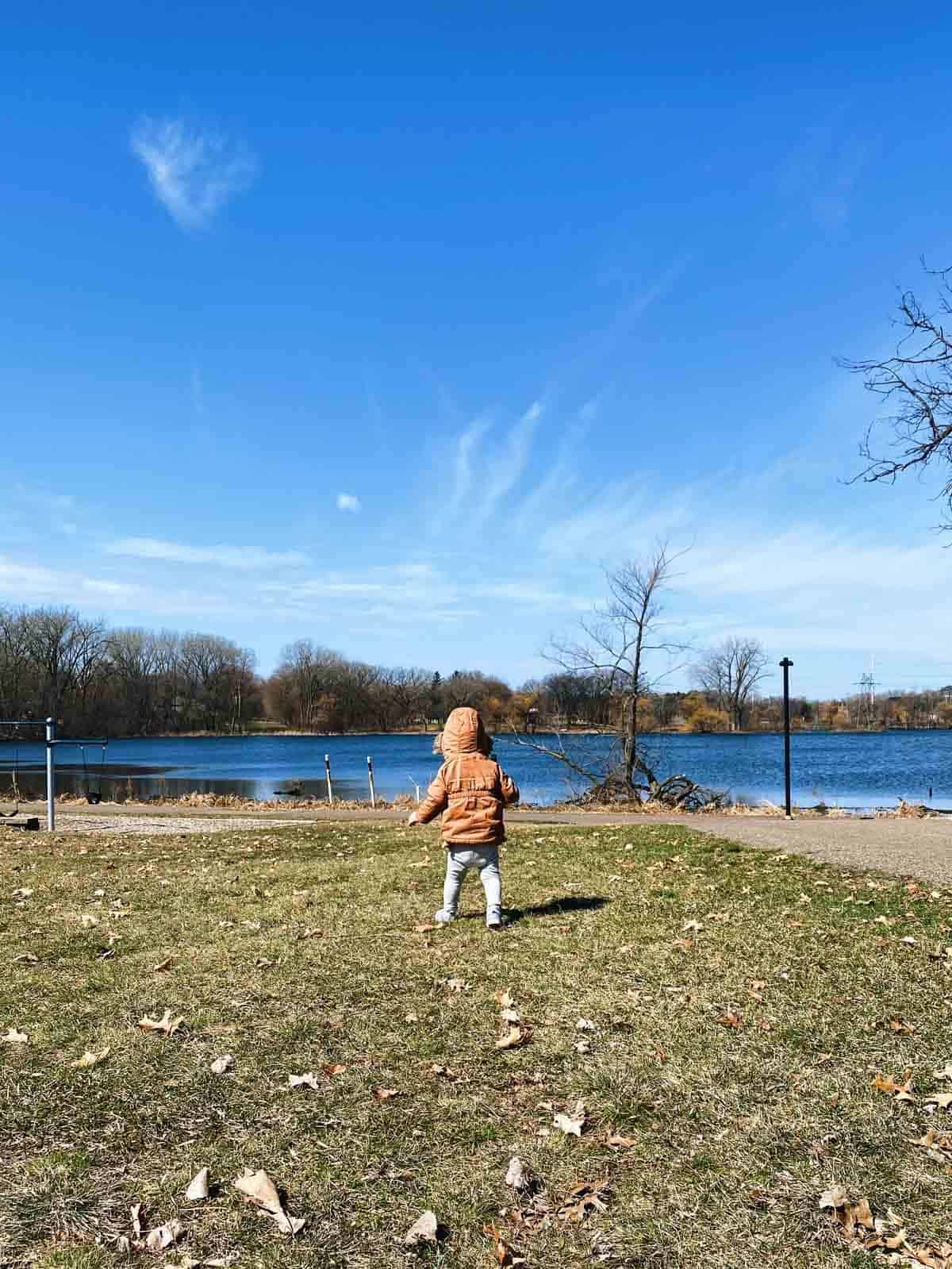 A young child in a brown jacket with the hood over their head walks near the water alone.