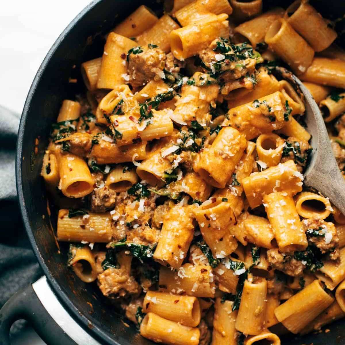 A bowl filled of Rigatoni with Sausage and Kale with a wooden spoon over it.