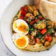 Creamy hummus topped with fresh herbs, sauteed cauliflower, tomatoes, eggs and pita in a bowl.