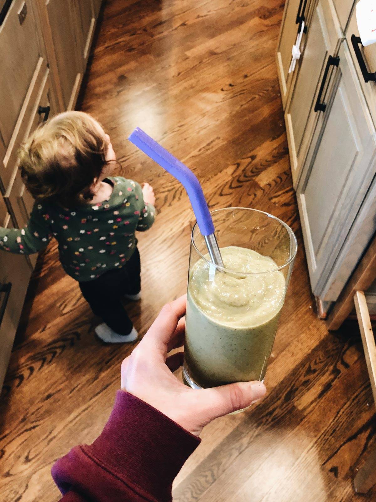 A kid is walking in the kitchen as a woman is holding a glass of smoothie with a straw in it.
