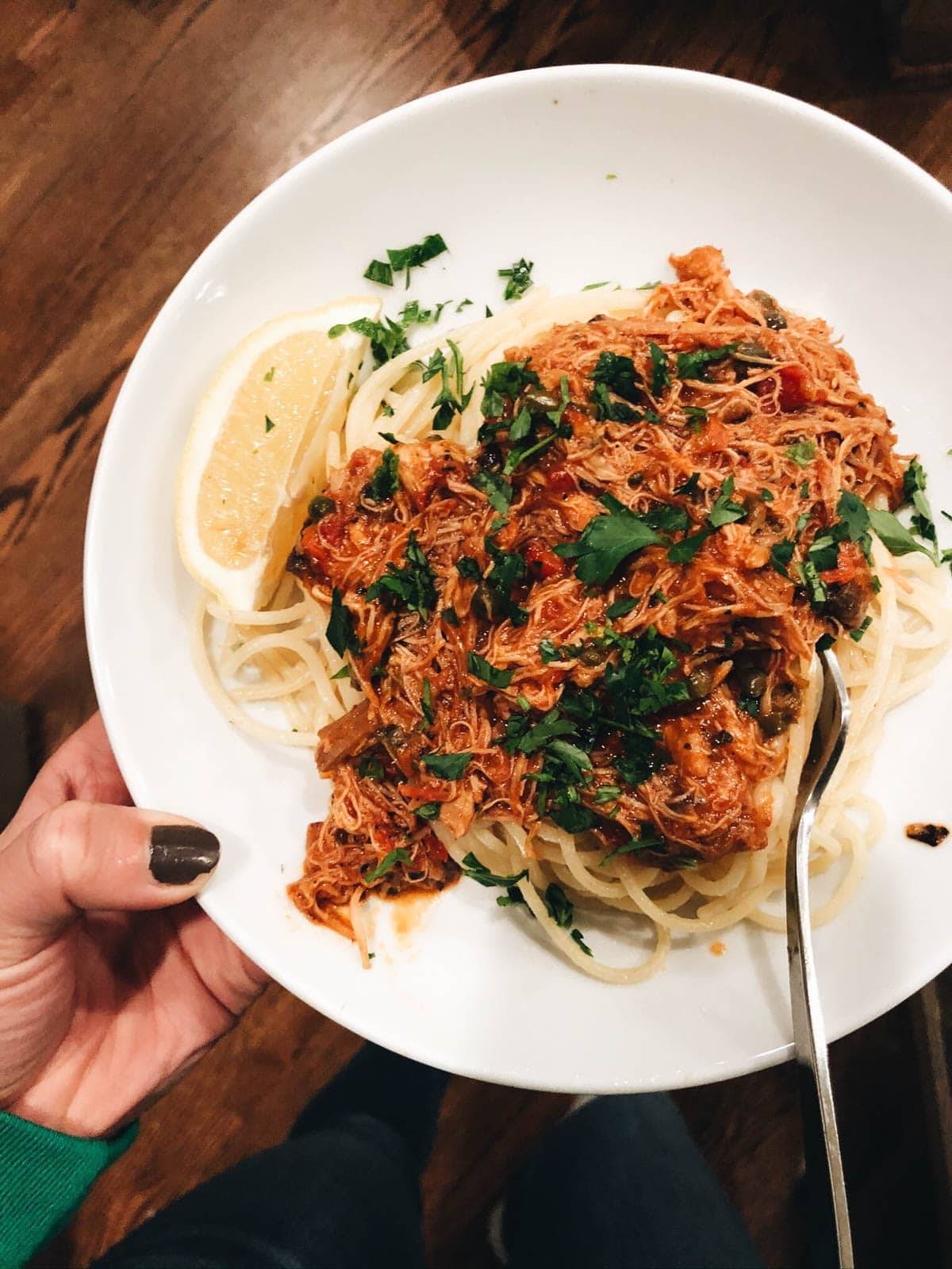 A big white bowl of spaghetti with a shredded chicken tomato sauce, topped with herbs and a lemon wedge.