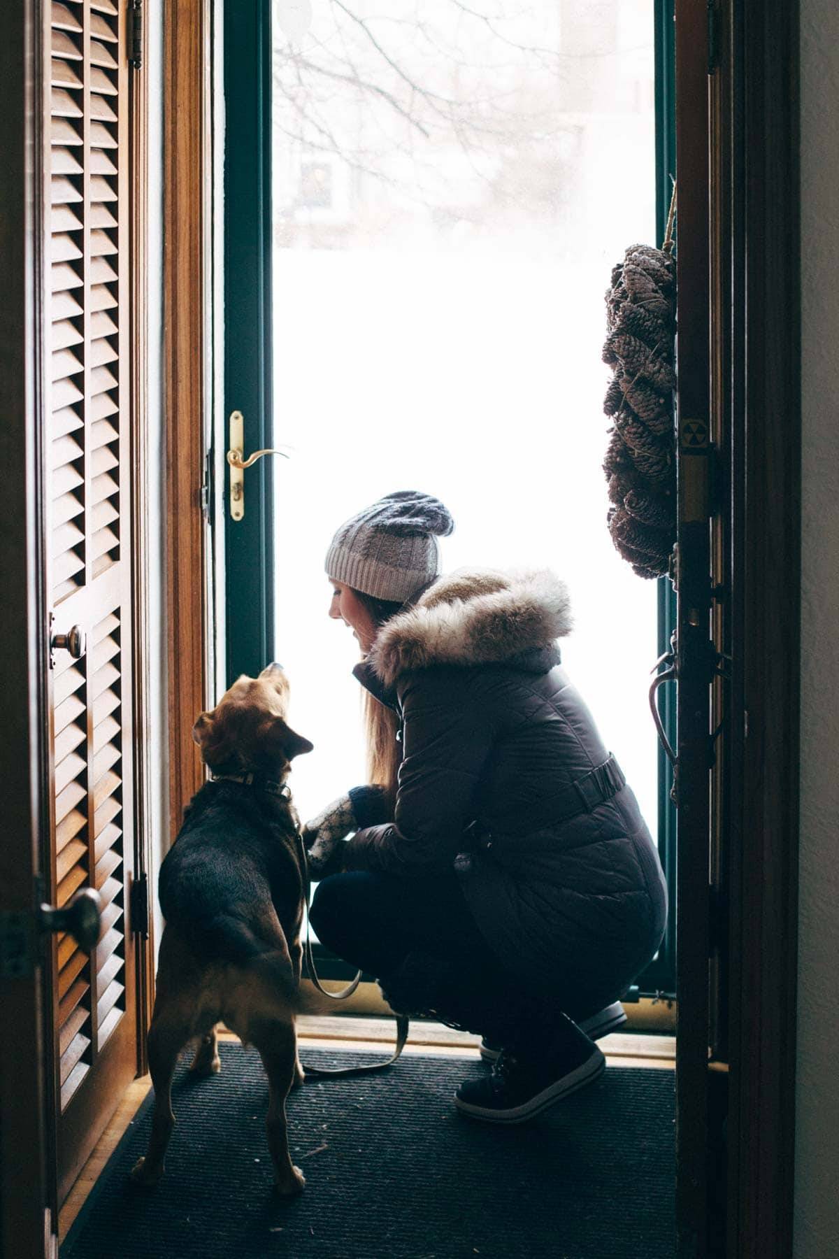 Woman dressed warmly kneeling next to a dog near a door looking out on a winter scene.