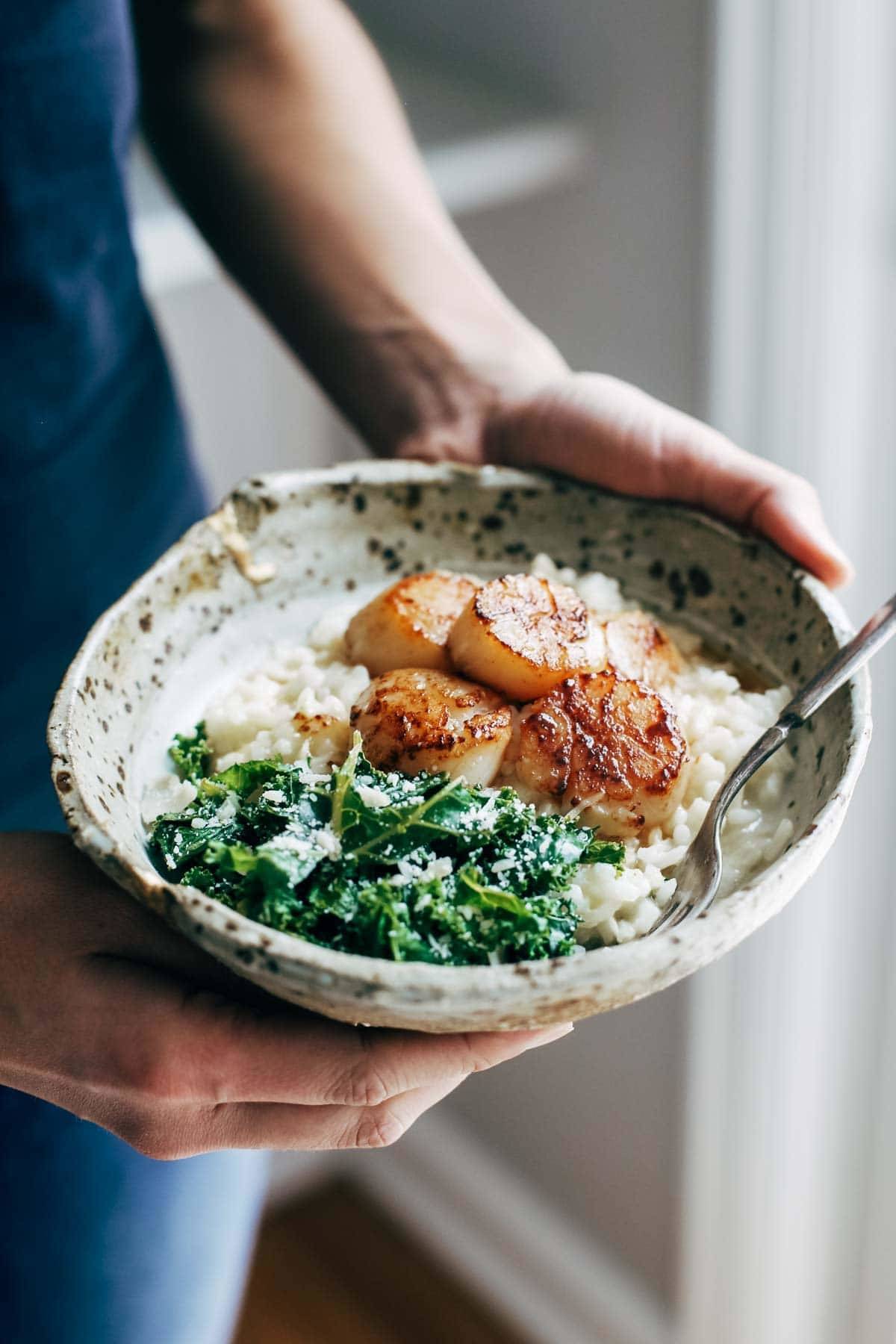 White hand holding a bowl with brown butter scallops, kale, and parmesan risotto. 