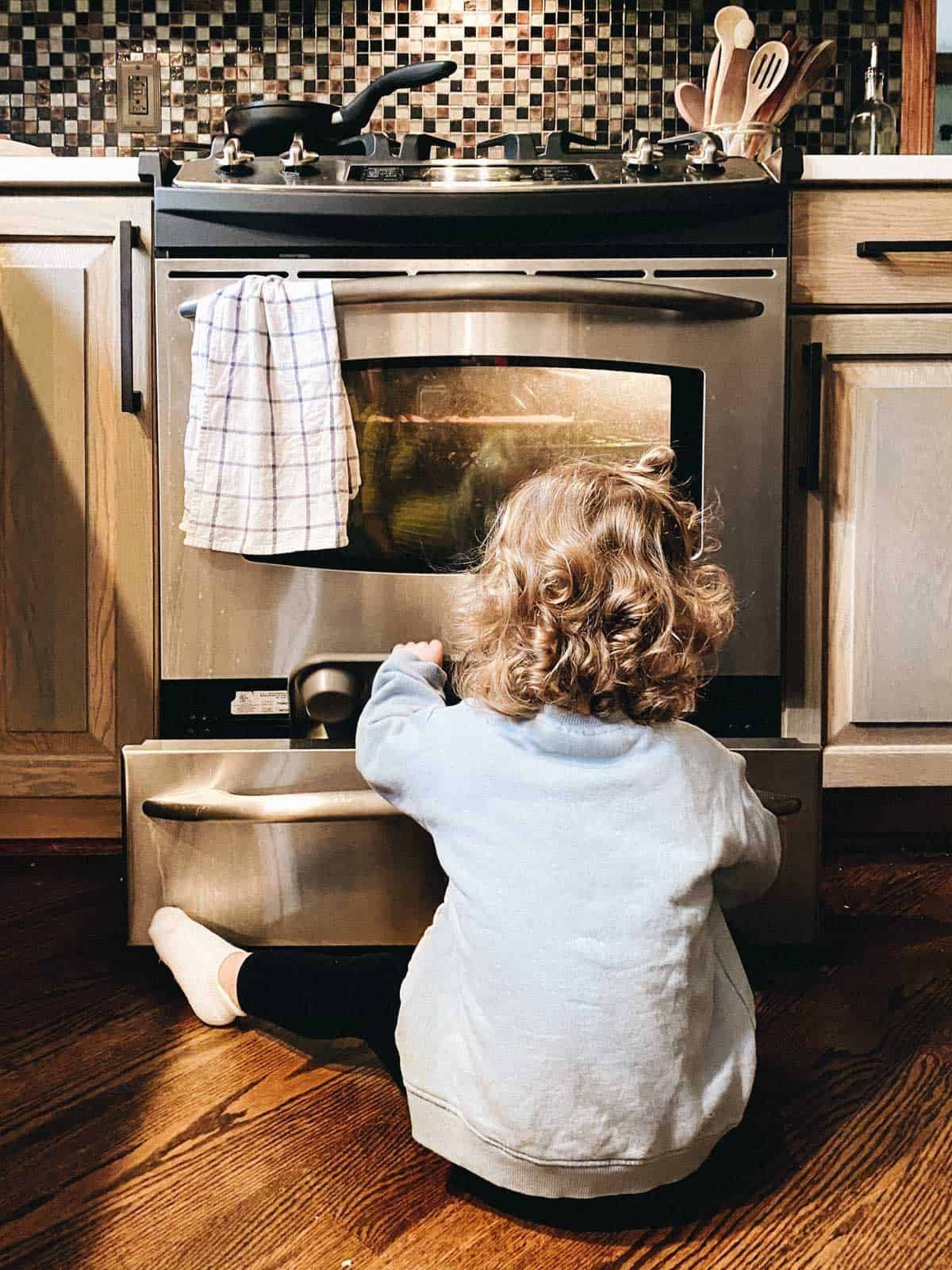 A child playing in the pullout storage area under an oven.