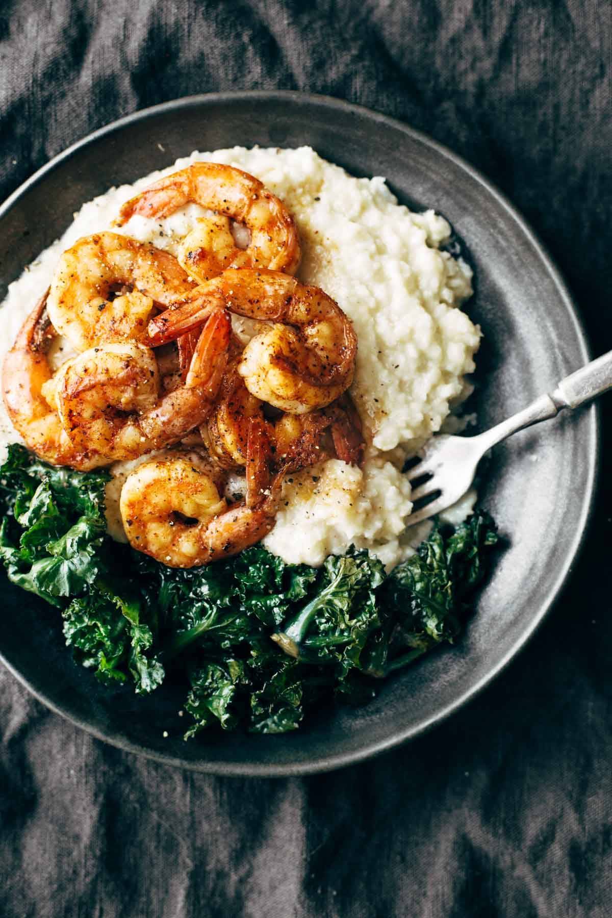 Perfectly cooked tail on shrimp with kale, and cauliflower mash in a large dark bowl.