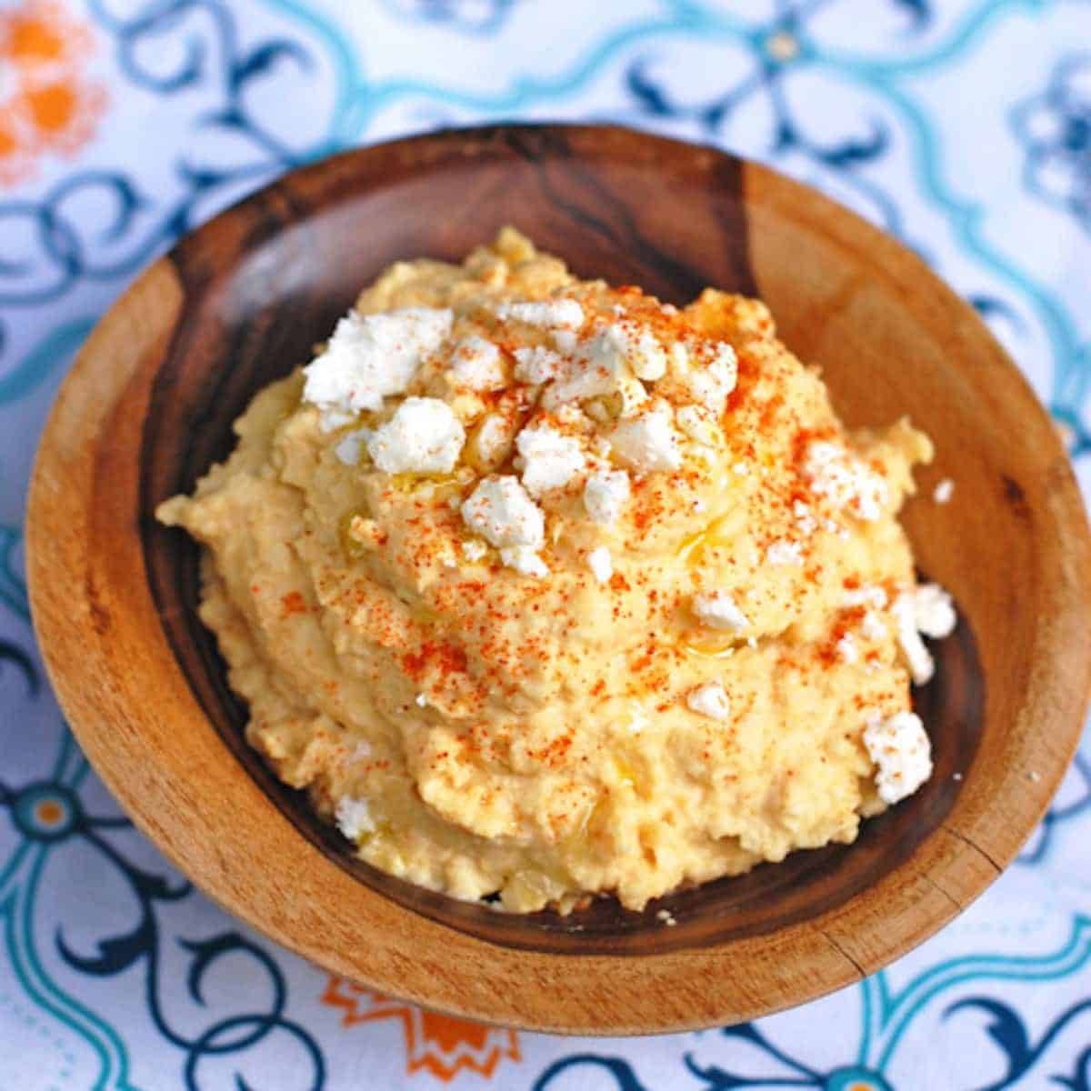 Spicy hummus topped with crushed red pepper, feta cheese, and olive oil in a wooden bowl.