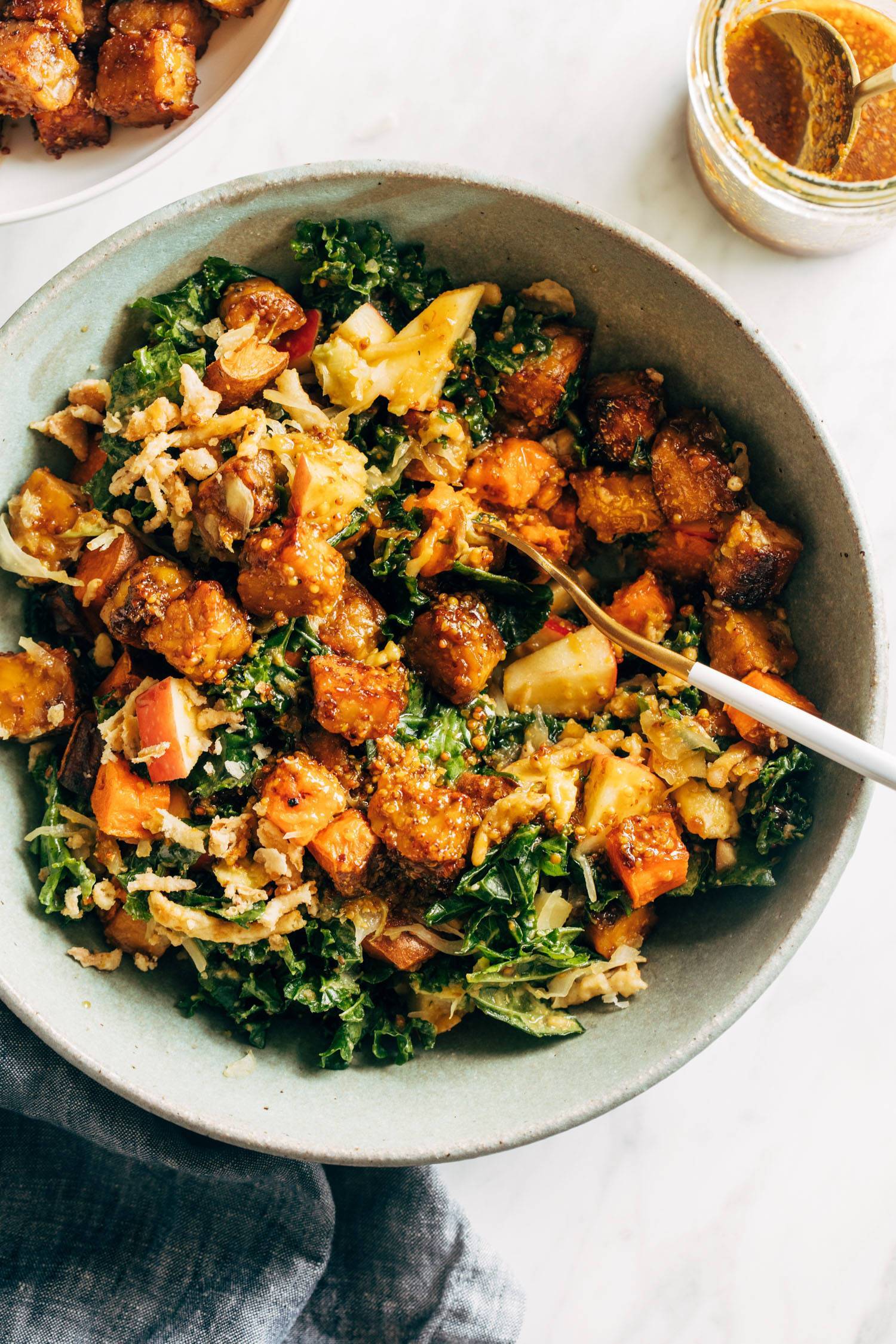 Maple mustard tempeh bowls in a grey bowl with a gold spoon. There's extra dressing and tempeh on the side and a blue napkin under the bowl. 