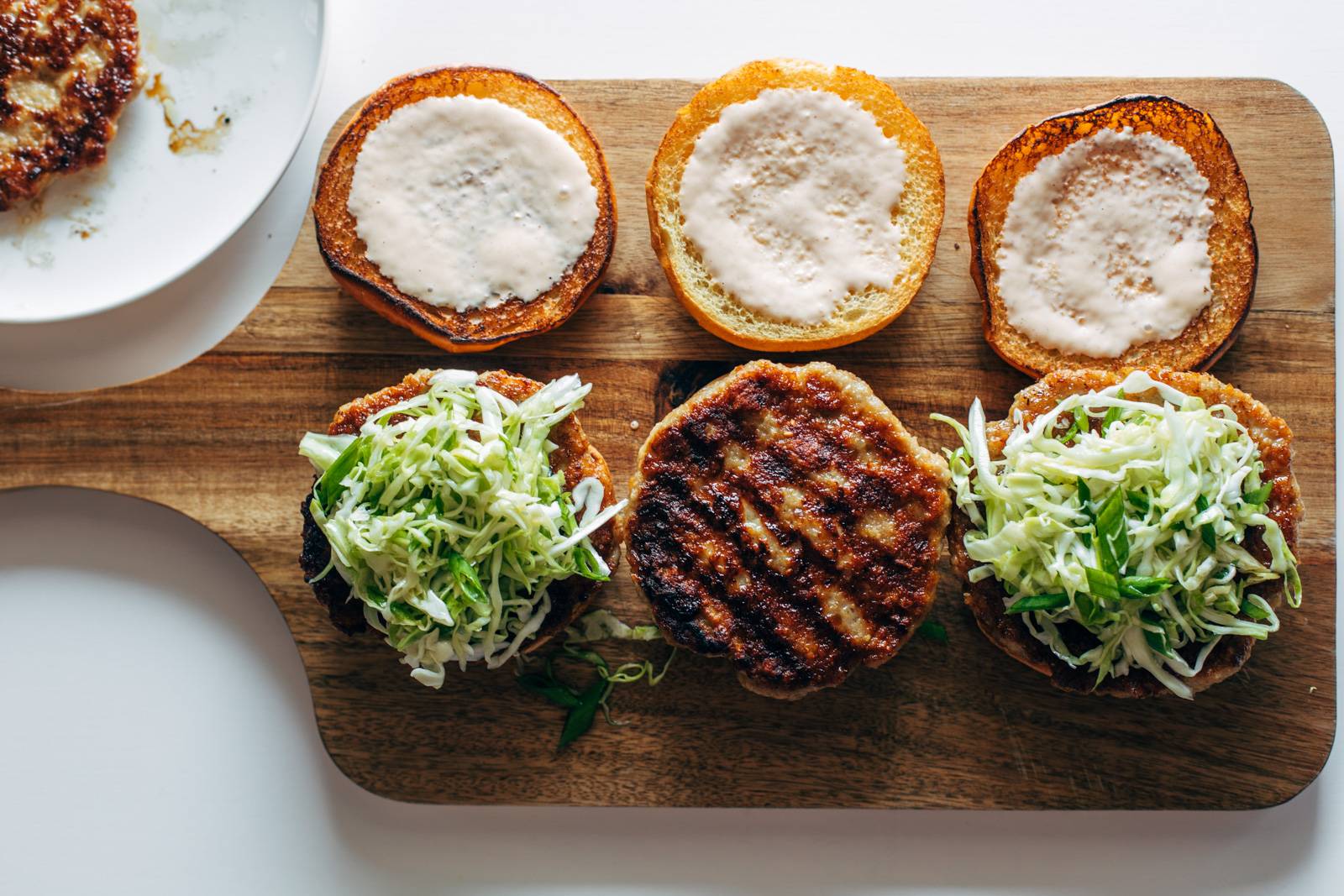 Assembling chicken burgers with buns, mayo, and slaw on a wooden serving board.