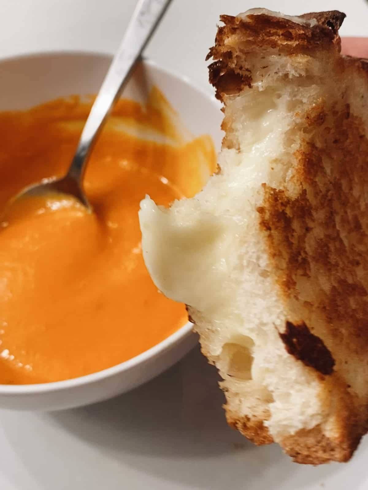 A bowl having tomato soup and spoon in it and a piece of bread lying near it.