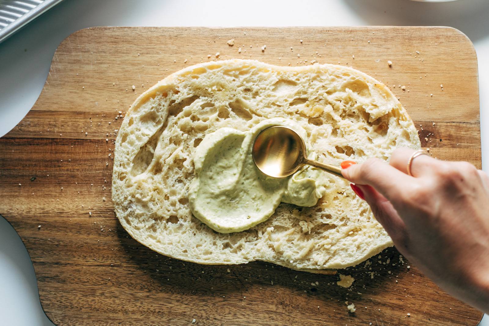White hand spreading a layer of pesto mayo on focaccia bread