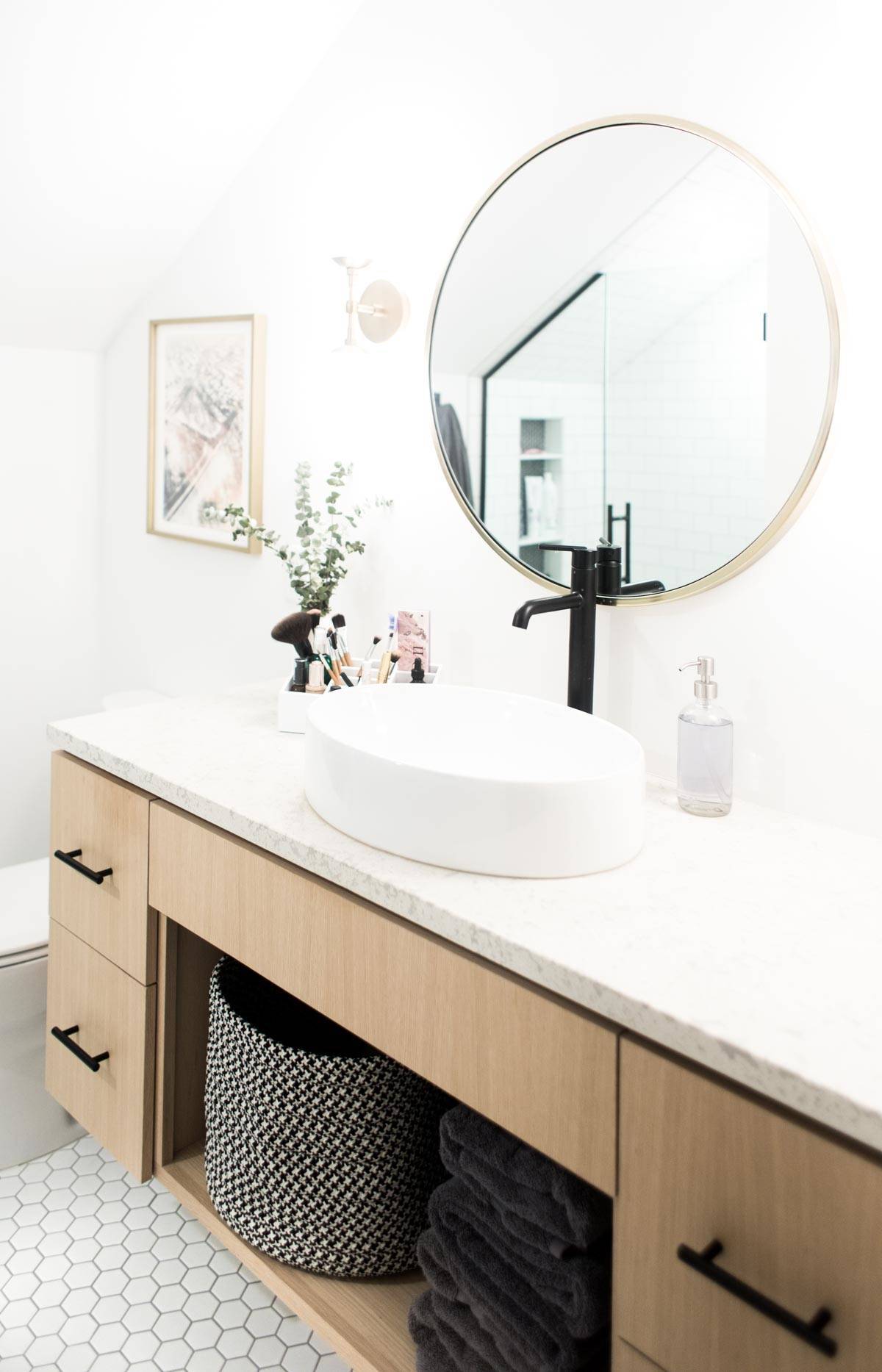 A modern white bathroom with a oval bowl sink on a long countertop and a round mirror above the sink.