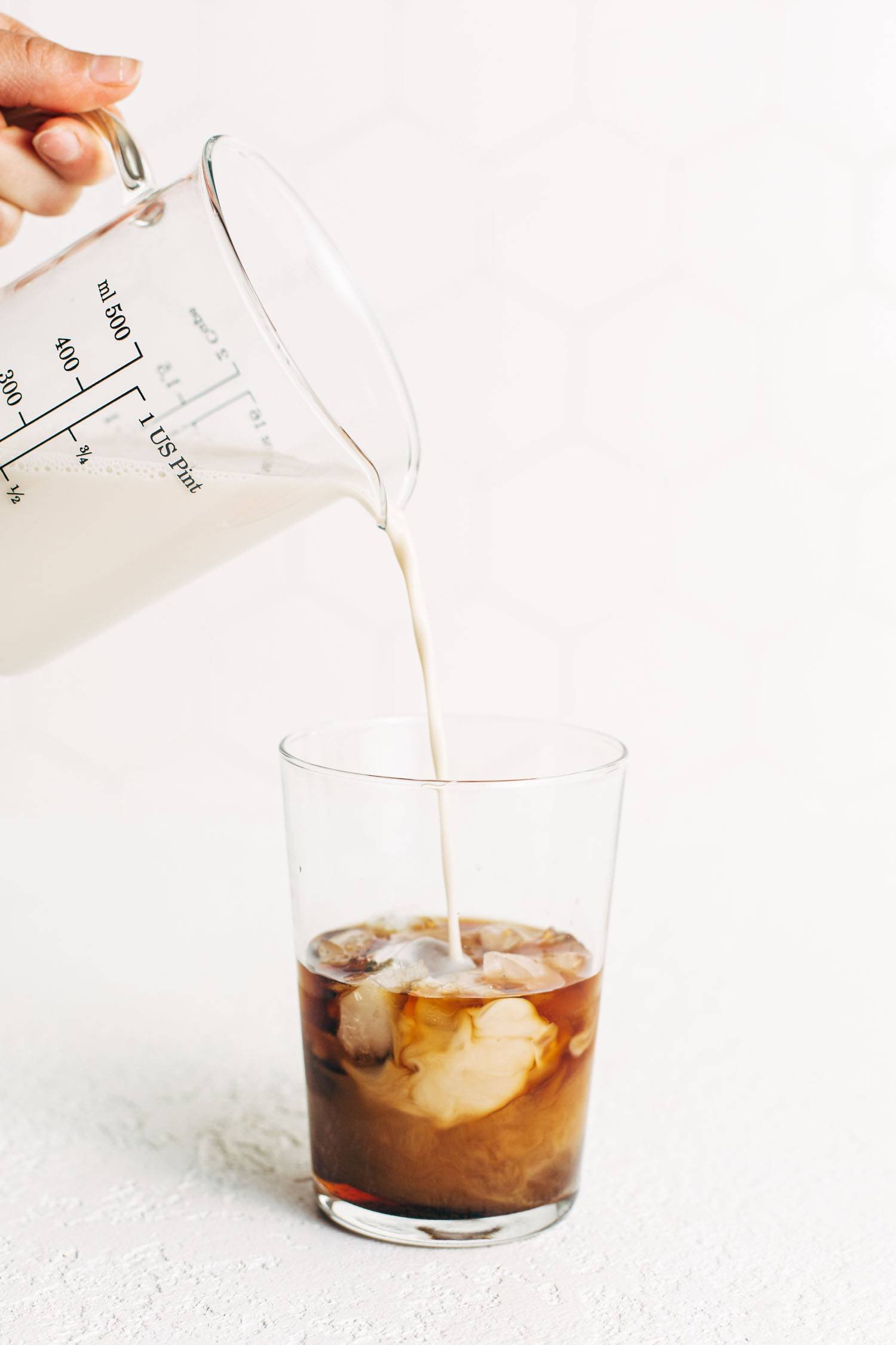 Measuring cup pouring oat milk into a glass of iced coffee.