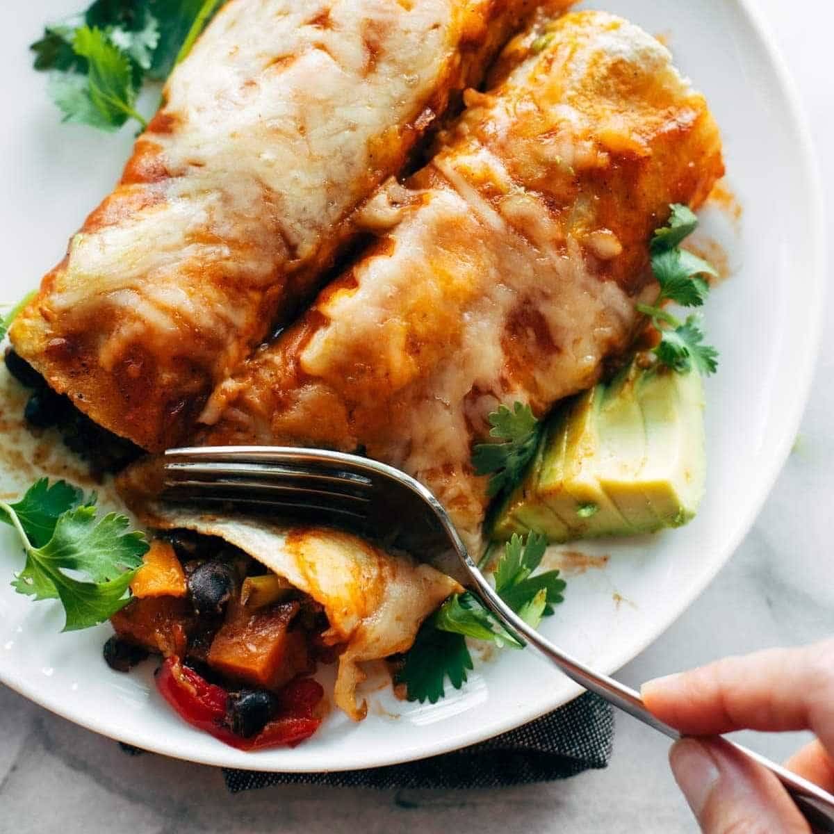 Person cutting an enchilada full of veggies and covered in sauce and cheese.