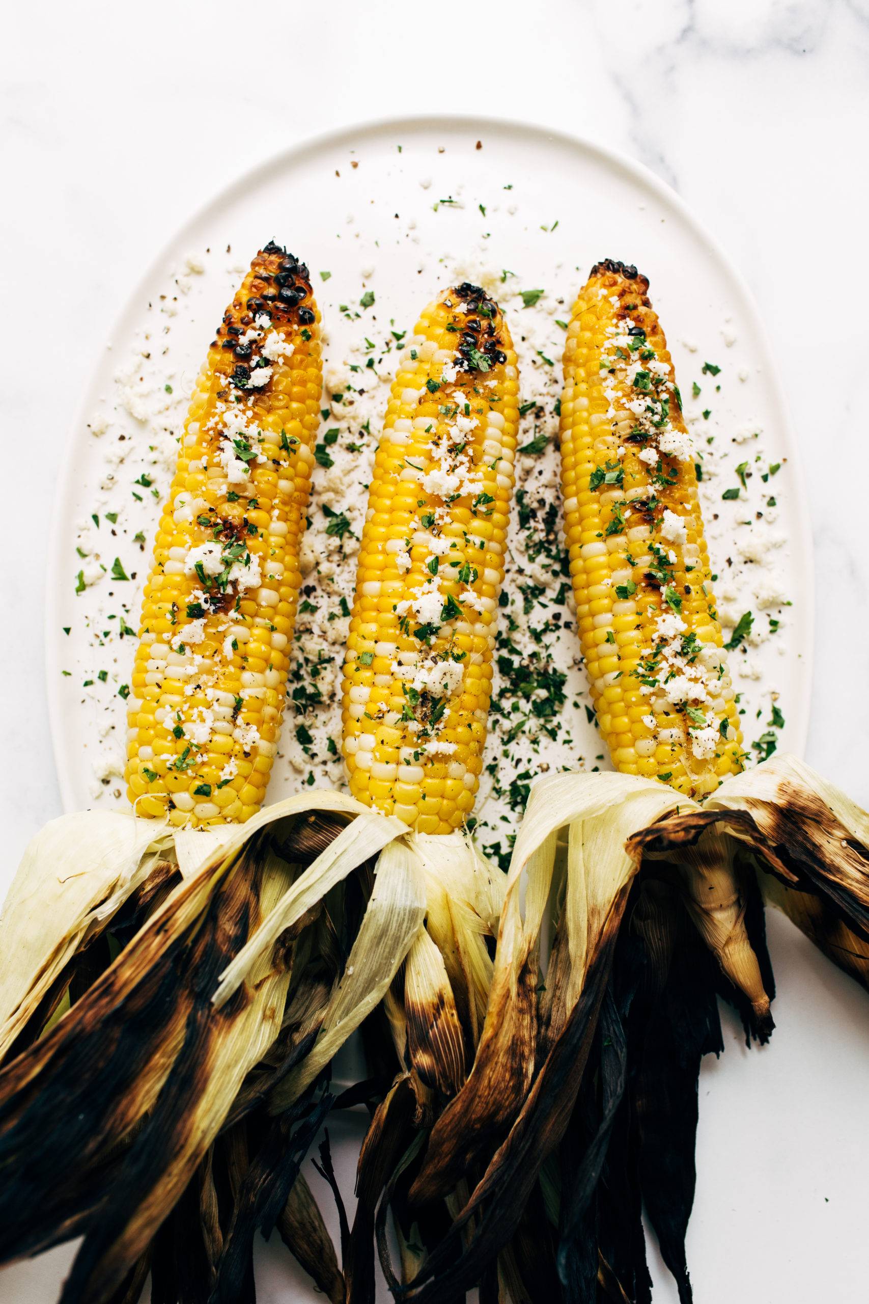 Three ears of grilled corn on a plate with husks pulled back, sprinkled with cheese and parsley.